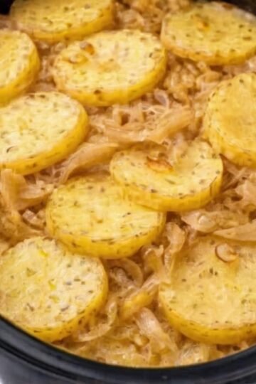 A rustic slow cooker potato and onion bake with tender golden potato slices layered over sweet caramelized onions, lightly coated in melted butter and seasoning, served hot in a black slow cooker.