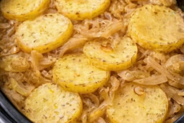 A rustic slow cooker potato and onion bake with tender golden potato slices layered over sweet caramelized onions, lightly coated in melted butter and seasoning, served hot in a black slow cooker.