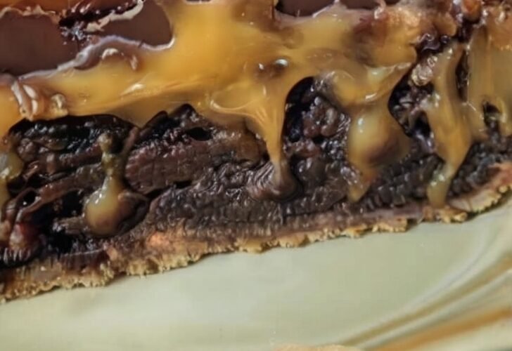 A rich caramel brownie pie slice with a buttery crust, fudgy chocolate brownie filling, gooey caramel layers, and a glossy chocolate topping.