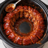 Alt text: Tender slow-cooked pork ribs coated in a thick, glossy barbecue sauce, arranged in a circle inside a black slow cooker, with a spoonful of rich sauce ready to drizzle over the juicy, flavorful meat.