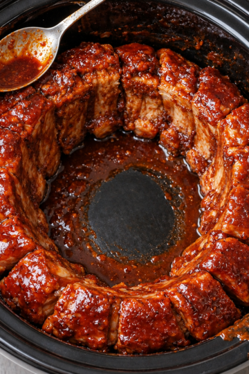 Alt text: Tender slow-cooked pork ribs coated in a thick, glossy barbecue sauce, arranged in a circle inside a black slow cooker, with a spoonful of rich sauce ready to drizzle over the juicy, flavorful meat.
