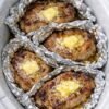 Alt text: Four golden-brown pork chops topped with melting butter and herbs, each wrapped in crinkled aluminum foil inside a white slow cooker, with savory juices pooling around the tender, juicy meat.