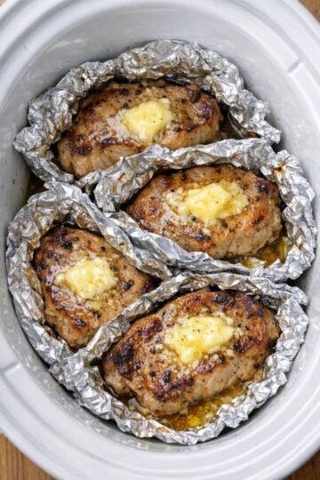 Alt text: Four golden-brown pork chops topped with melting butter and herbs, each wrapped in crinkled aluminum foil inside a white slow cooker, with savory juices pooling around the tender, juicy meat.