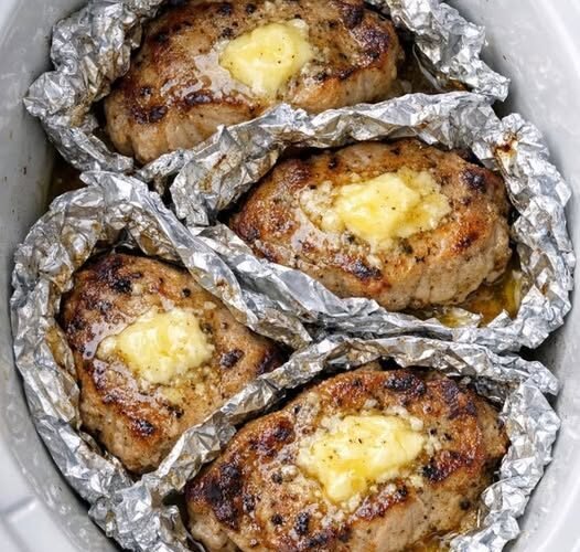 Alt text: Four golden-brown pork chops topped with melting butter and herbs, each wrapped in crinkled aluminum foil inside a white slow cooker, with savory juices pooling around the tender, juicy meat.