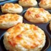 A batch of fluffy homemade cheddar biscuits with golden, crispy cheese-covered tops and soft, tender centers, freshly baked in a muffin-style pan.