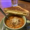 A cozy bowl of hearty tomato-lentil soup topped with creamy cheese and black pepper, served alongside a perfectly toasted grilled cheese sandwich with a golden crust and gooey melted cheese.