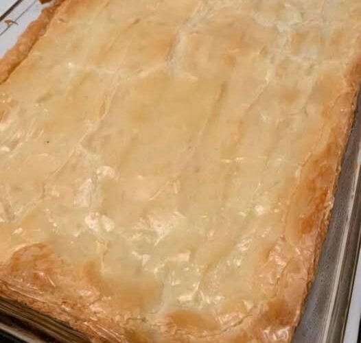 A golden, flaky pastry-topped casserole baked in a rectangular glass dish, with a beautifully crisp, buttery crust and lightly browned edges. simplyhappenings.com