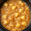 Tender chunks of potatoes and sliced onions simmering in a savory, golden-brown gravy in a black slow cooker, creating a hearty, comforting side dish with a rich, flavorful sauce.