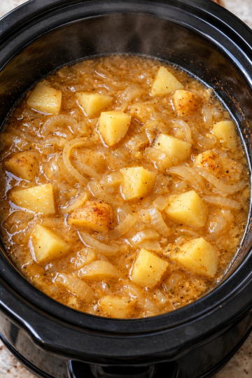 Tender chunks of potatoes and sliced onions simmering in a savory, golden-brown gravy in a black slow cooker, creating a hearty, comforting side dish with a rich, flavorful sauce.