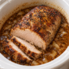 Alt text: Juicy slow-cooked pork roast with a golden-brown, herb-seasoned crust, sliced to reveal tender meat, surrounded by rich brown gravy and soft onions in a white slow cooker.